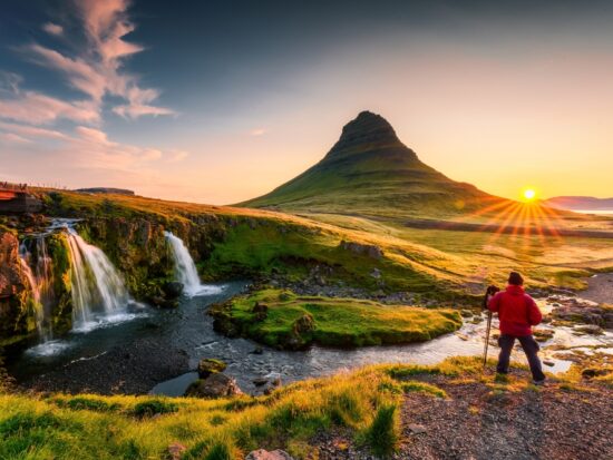 person wearing a red jacket and black pants stands in front of Kirkjufell mountain in iceland and there is a waterfall to the left and sunset to the right