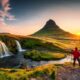 person wearing a red jacket and black pants stands in front of Kirkjufell mountain in iceland and there is a waterfall to the left and sunset to the right