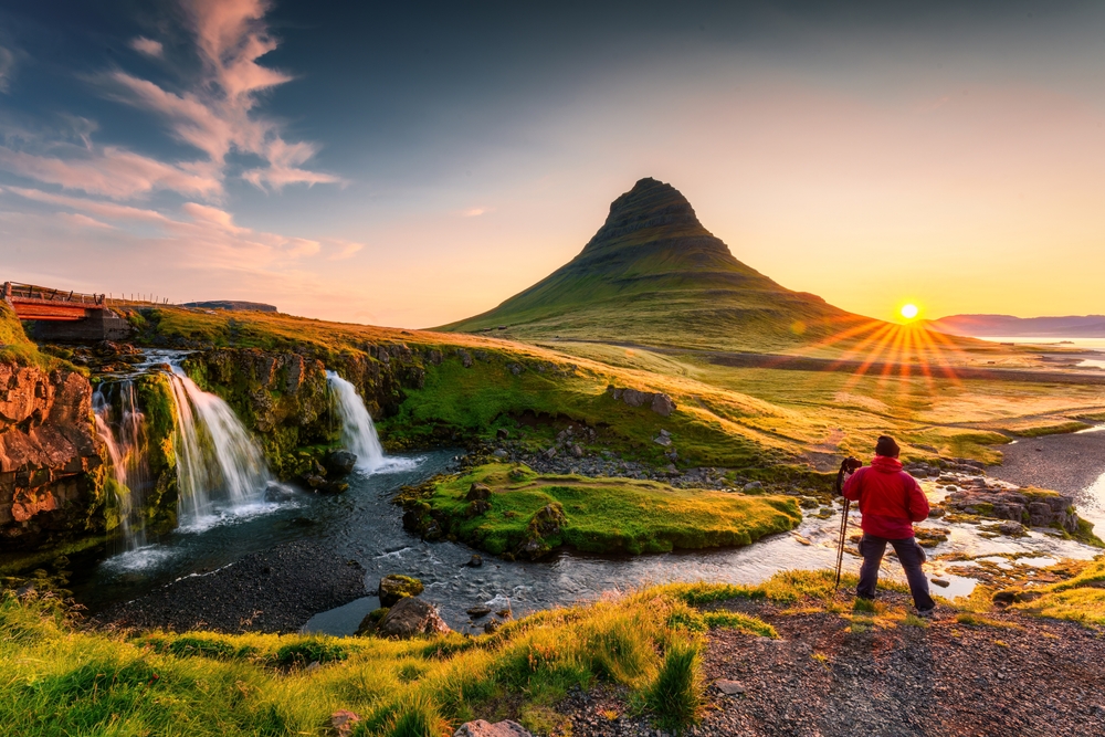 A young tourist walks down a winding road as the green of summer in Iceland fades: she is well prepared and knows what to wear to Iceland in the fall with her knit beanie, hiking pants, boots, and parka with a fur hood.