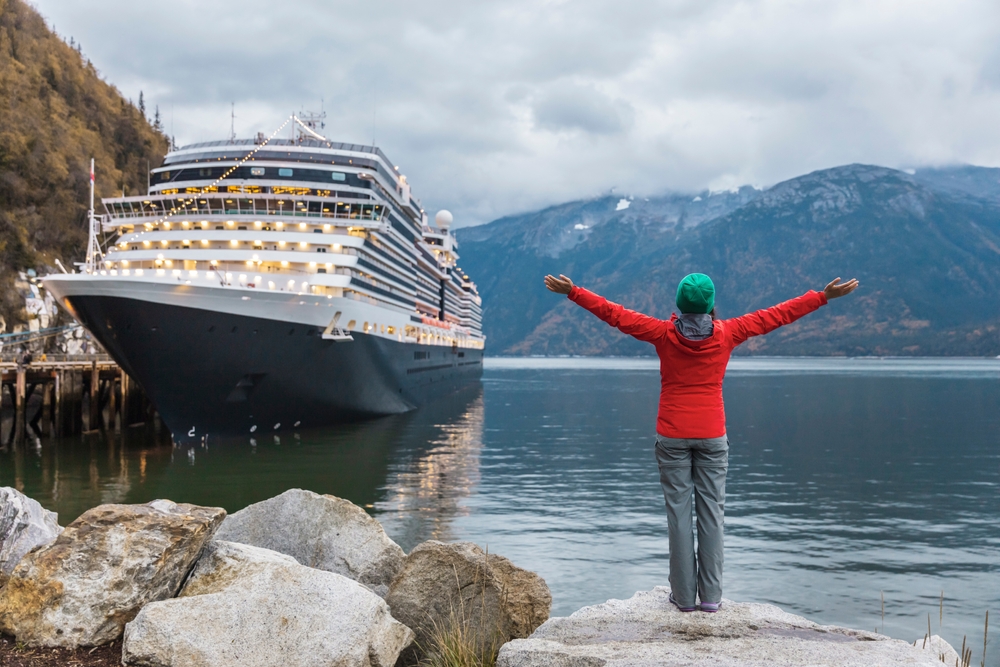 A woman stands with her hands spread wide, excited as a cruise ship docks: she wears a green beanie, orange jacket, and rain pants.