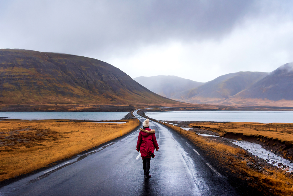 A young tourist walks down a winding road as the green of summer in Iceland fades: she is well prepared and knows what to wear to Iceland in the fall with her knit beanie, hiking pants, boots, and parka with a fur hood.