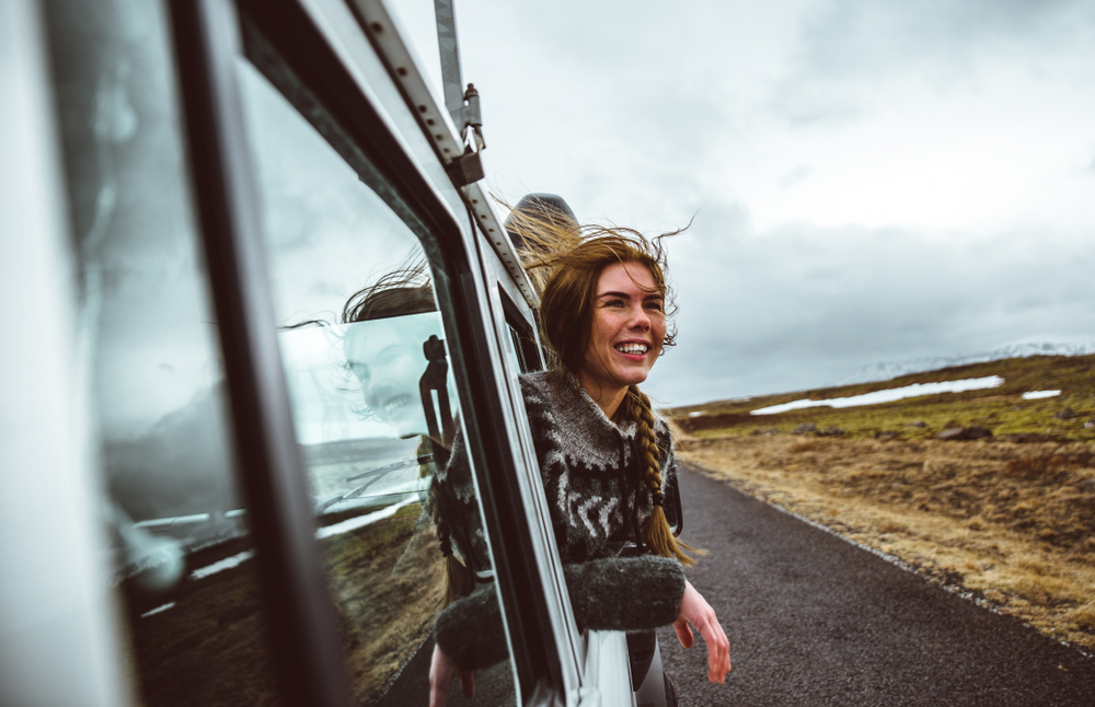 A woman sticks her head out the window of a car as they drive down a road, her hair is in a braid and she wears a wool sweater, perfect for road trips since she knew what to wear to Iceland in the fall.