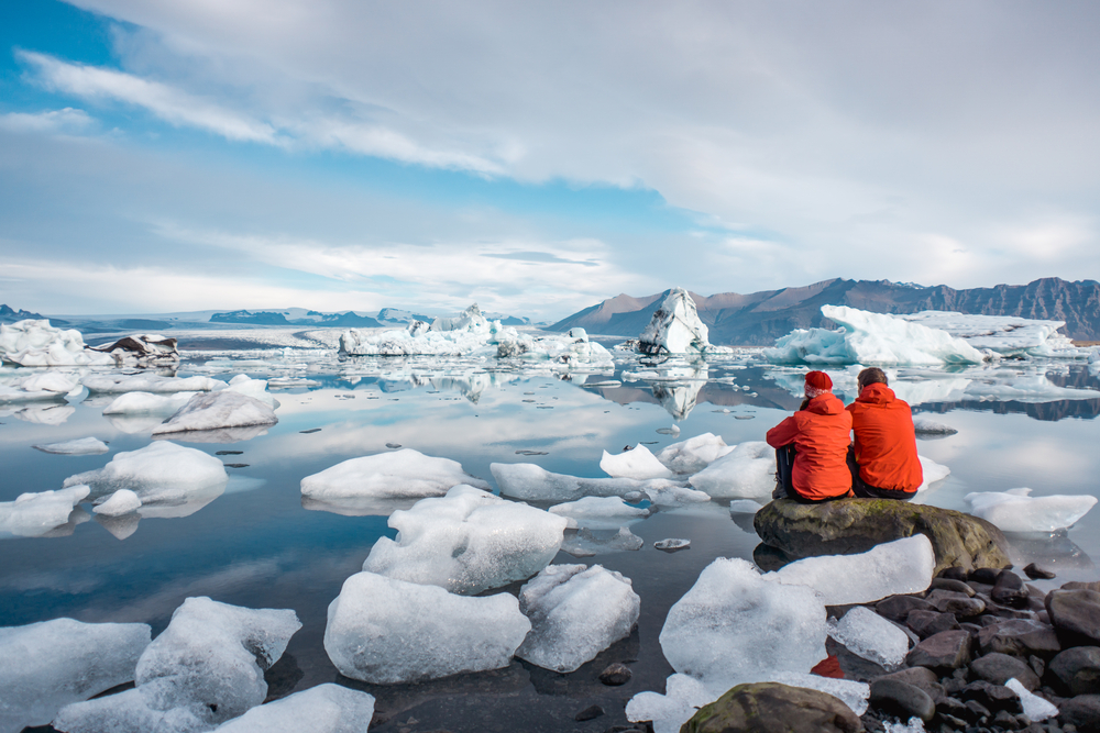 A couple sits on the edge of a rock, overlooking ice formations on the coast: they knew what to wear to Iceland in the fall with their hats and matching orange parkas.