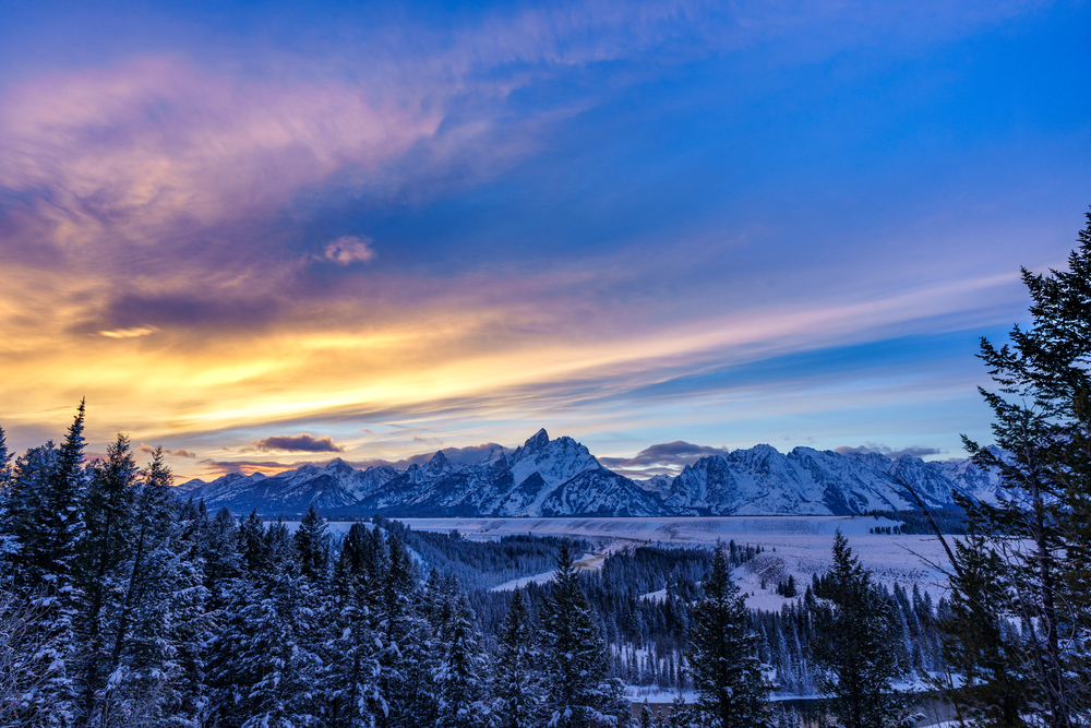 During a bright sunrise, the sun stretches over the snowy mountains and trees in Jackson Hole, one of the Best Destinations For A Magical White Christmas In The USA,