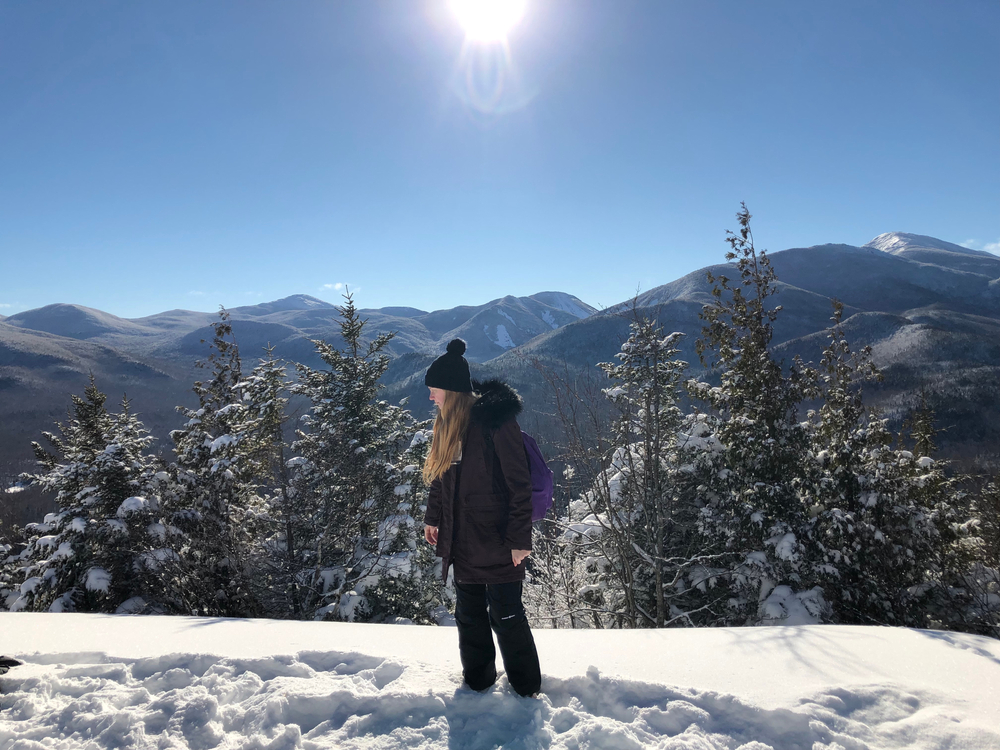 A woman walks through snow in Lake Placid, a parka and hoodie and beanie on.