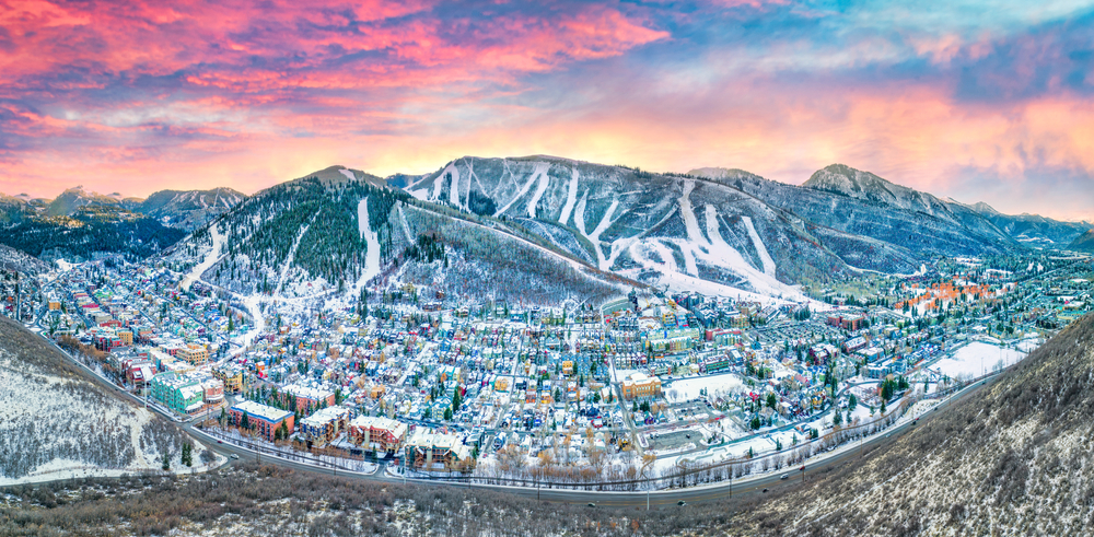 Some the of Best Destinations For A Magical White Christmas In The USA feature mountain towns, as seen in this photo, with homes on the bottom of hills, snow gathering in a pink sky.