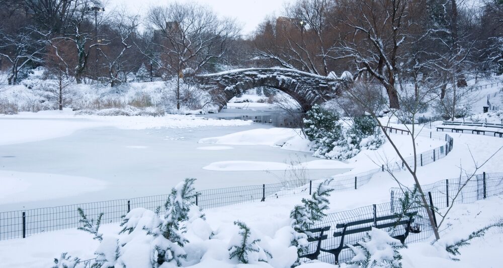 In Park City, which is one of the Best Destinations For A Magical White Christmas In The USA, a little park with benches, a lake and bridge are covered in snow.