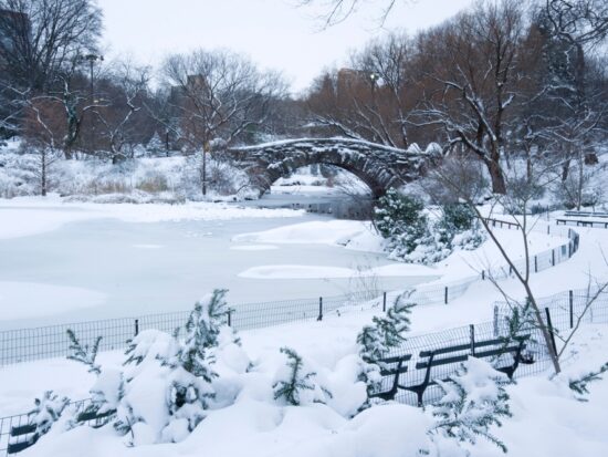 In Park City, which is one of the Best Destinations For A Magical White Christmas In The USA, a little park with benches, a lake and bridge are covered in snow.