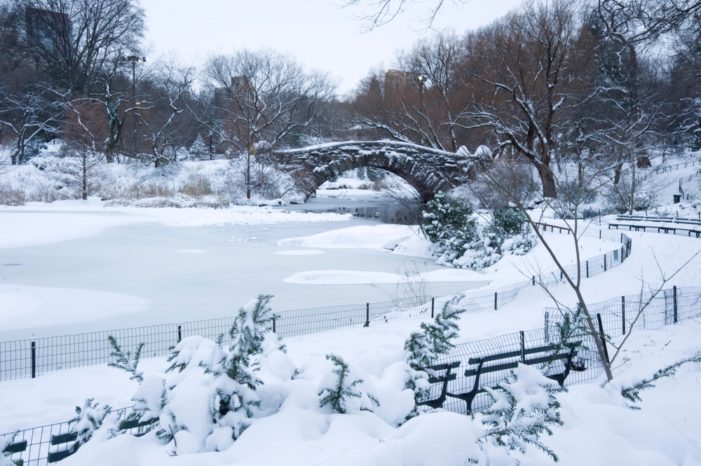 In Park City, which is one of the Best Destinations For A Magical White Christmas In The USA, a little park with benches, a lake and bridge are covered in snow.