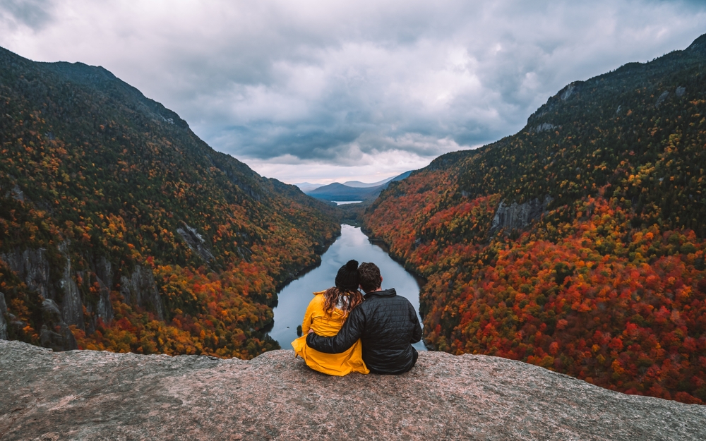 A couple sits on a rock formation, overlooking a river and the trees as they turn colors.