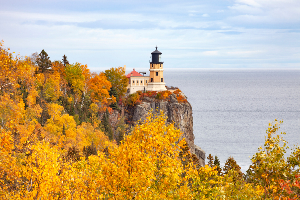 In Minnesota a photo shows a coast of water, a lighthouse on a ledge with yellow and orange foliage around it.