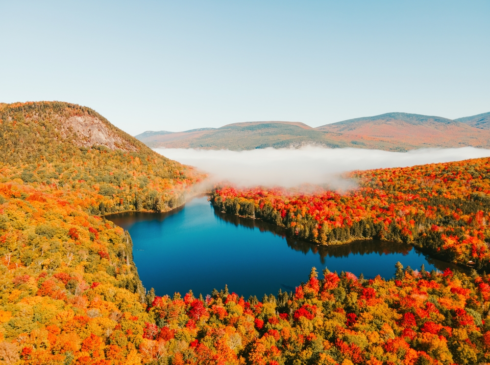 An overhead shot of fall foliage in New Hampshire shows trees turning colors as mist rives above hills and water.