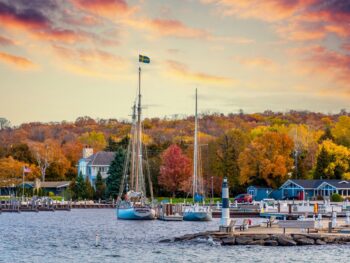 In Wisconsin, one of the Best States for Fall Foliage in the USA, boats sit on a lake at sunset, with tons of fall foliage in the backdrop.