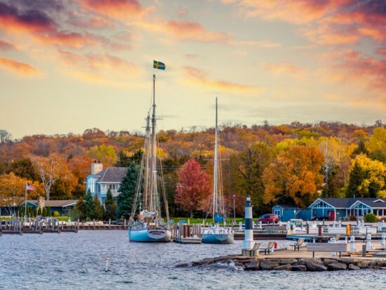 In Wisconsin, one of the Best States for Fall Foliage in the USA, boats sit on a lake at sunset, with tons of fall foliage in the backdrop.
