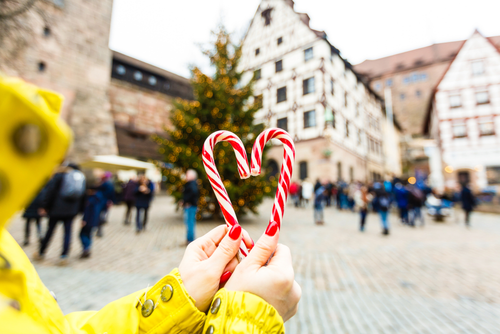 Two hands with red nails and a yellow jacket hold candy canes together in a town square, and they are enjoying time, which is one of the main Christmas travel tips for a smoother winter vacation.