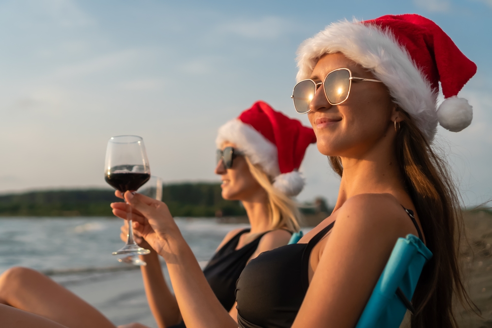 Two girls sit back on the beach enjoying glasses of wine during golden hour, sunglasses on and Santa hats prominent.