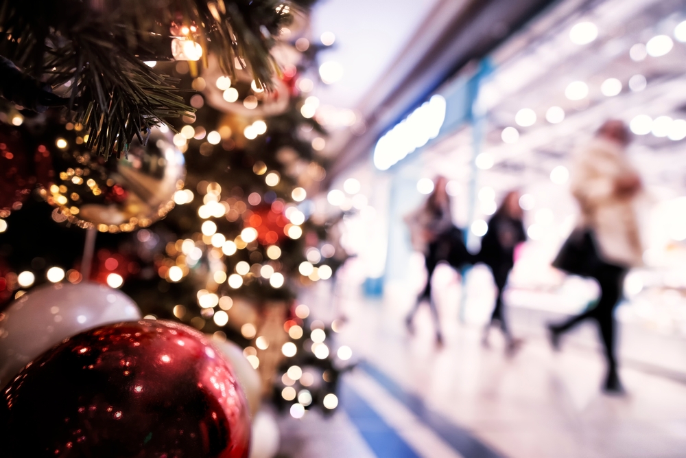 Guests rush by in a mall, blurred in the background, as a lit Christmas tree takes up the front foreground: slow down and enjoy the time is one of the best Christmas travel tips for a smoother winter vacation.