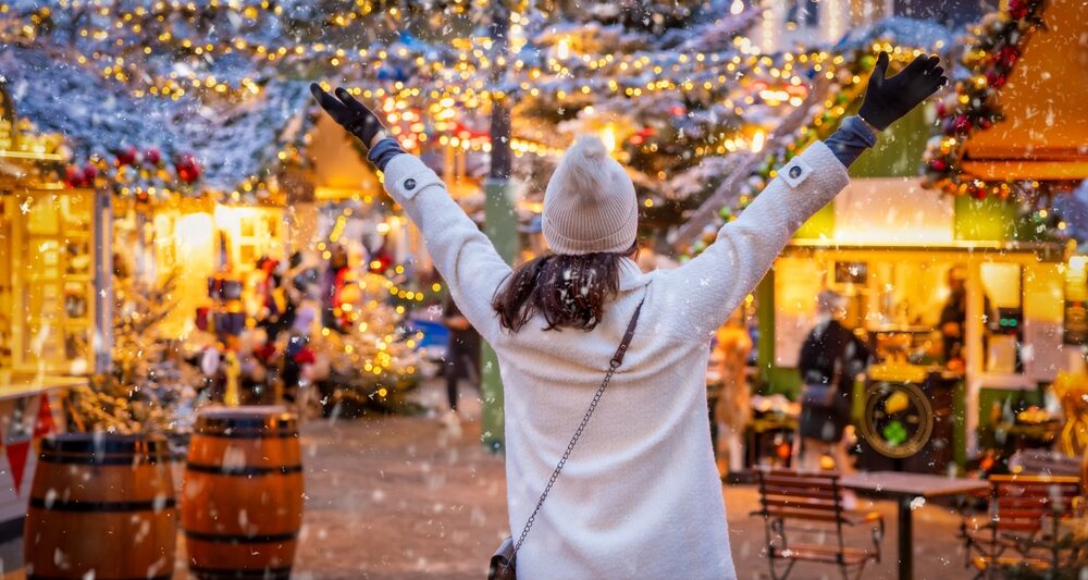 woman in white coat and white hat holding her arms out in joy, she is wearing black gloves and a crossbody purse. she is at a christmas market and you can see snow and the winter lights around her