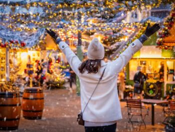 woman in white coat and white hat holding her arms out in joy, she is wearing black gloves and a crossbody purse. she is at a christmas market and you can see snow and the winter lights around her