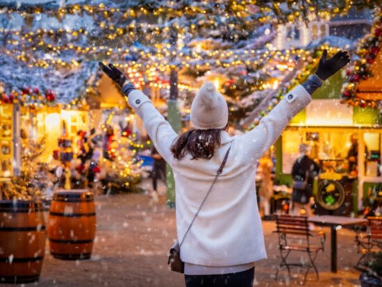 woman in white coat and white hat holding her arms out in joy, she is wearing black gloves and a crossbody purse. she is at a christmas market and you can see snow and the winter lights around her