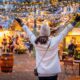 woman in white coat and white hat holding her arms out in joy, she is wearing black gloves and a crossbody purse. she is at a christmas market and you can see snow and the winter lights around her