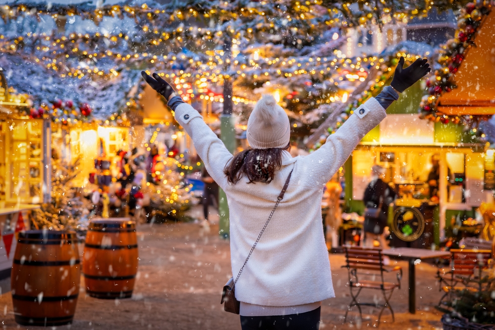 woman in white coat and white hat holding her arms out in joy, she is wearing black gloves and a crossbody purse. she is at a christmas market and you can see snow and the winter lights around her