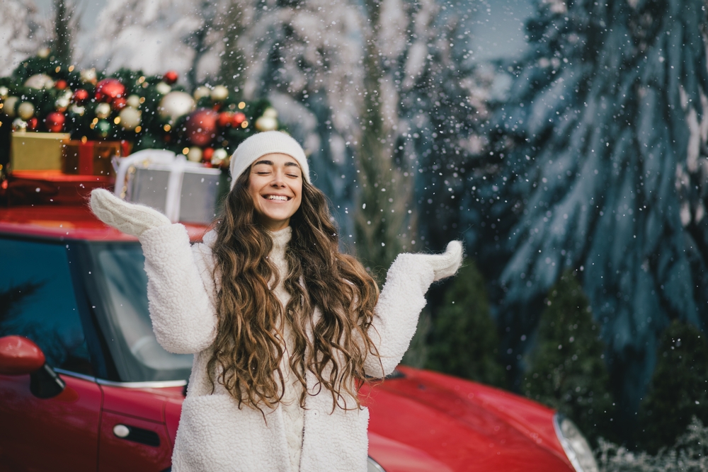 A woman poses in front of a car that is loaded with presents, snow falling around her, in her white hat, gloves and jacket.