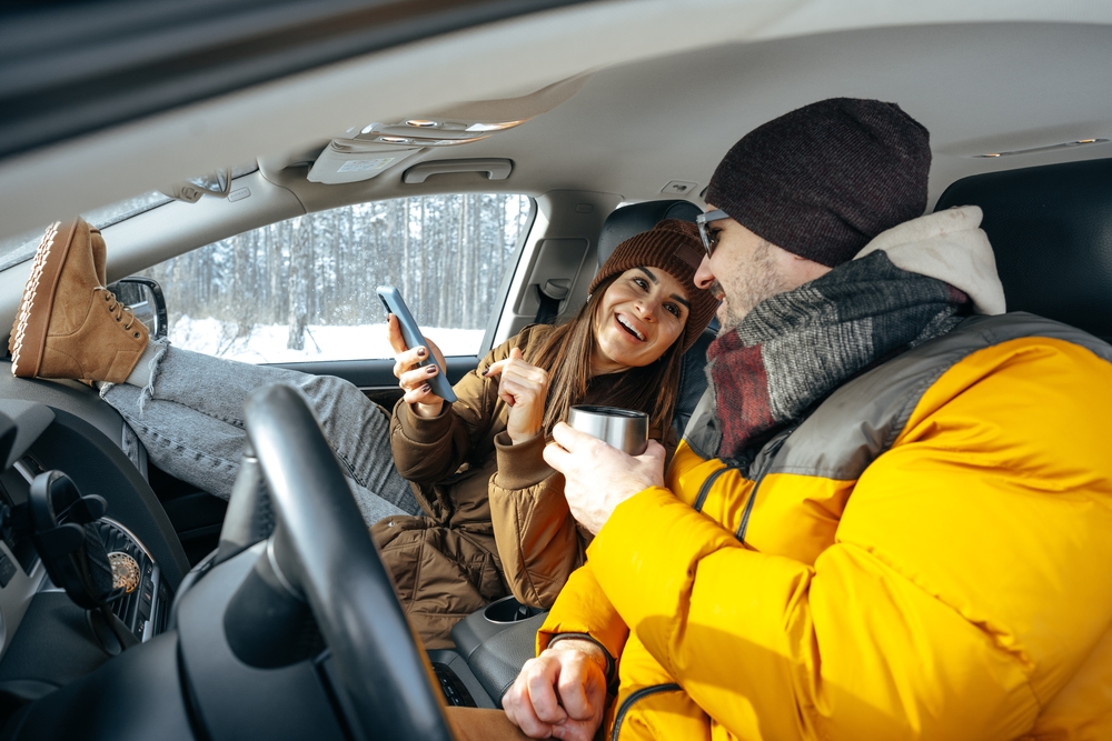 A couple lounges in a car, layered up with puffer jackets and hats and boots (layers are a main Christmas travel tips for a smoother winter vacation!) they smile at each other.