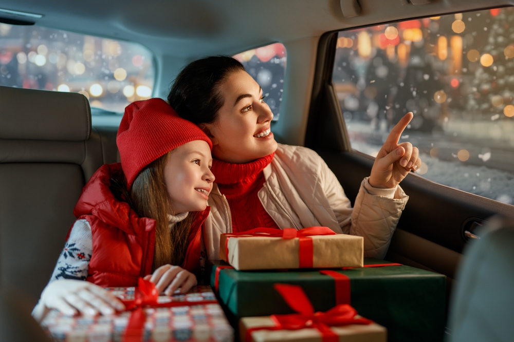 A little girl leans into her mothers shoulder, presents on their laps in the back of a car. They look out the window and point at the Christmas lights.