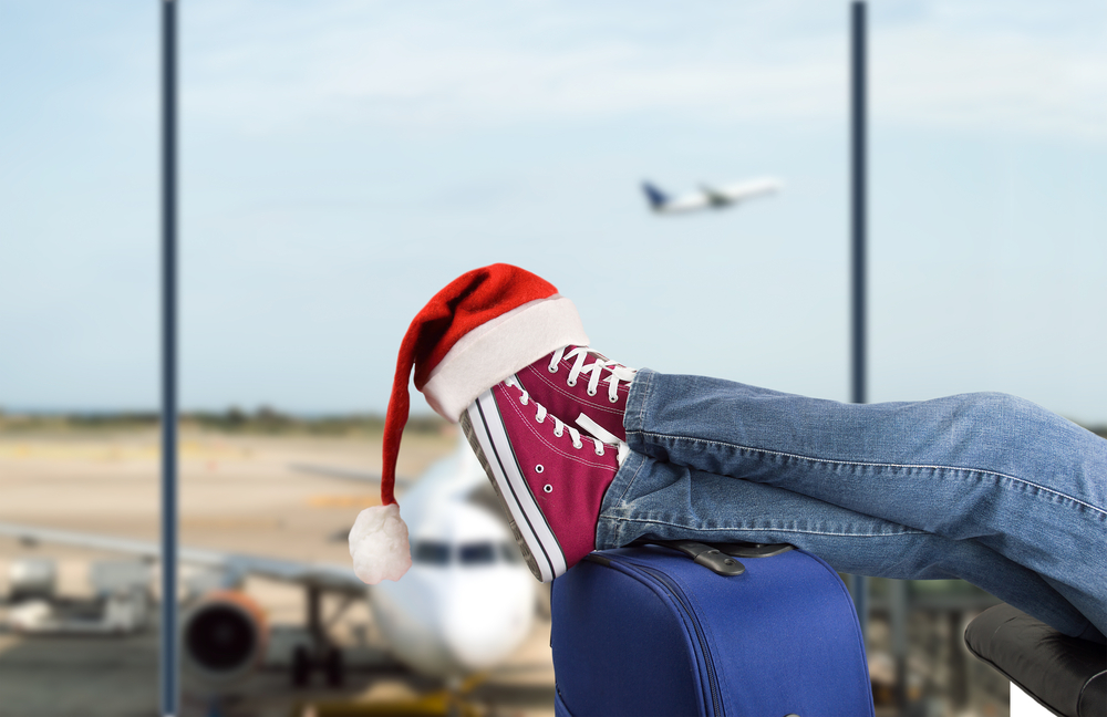 A person kicks back with their shoes and legs over their suitcase, a santa hat hanging off their toes.