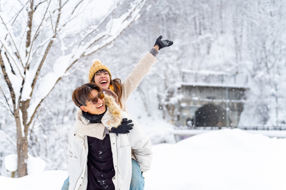 A girl jumps on a boys back and throws her hands up in the middle of a winter wonderland.