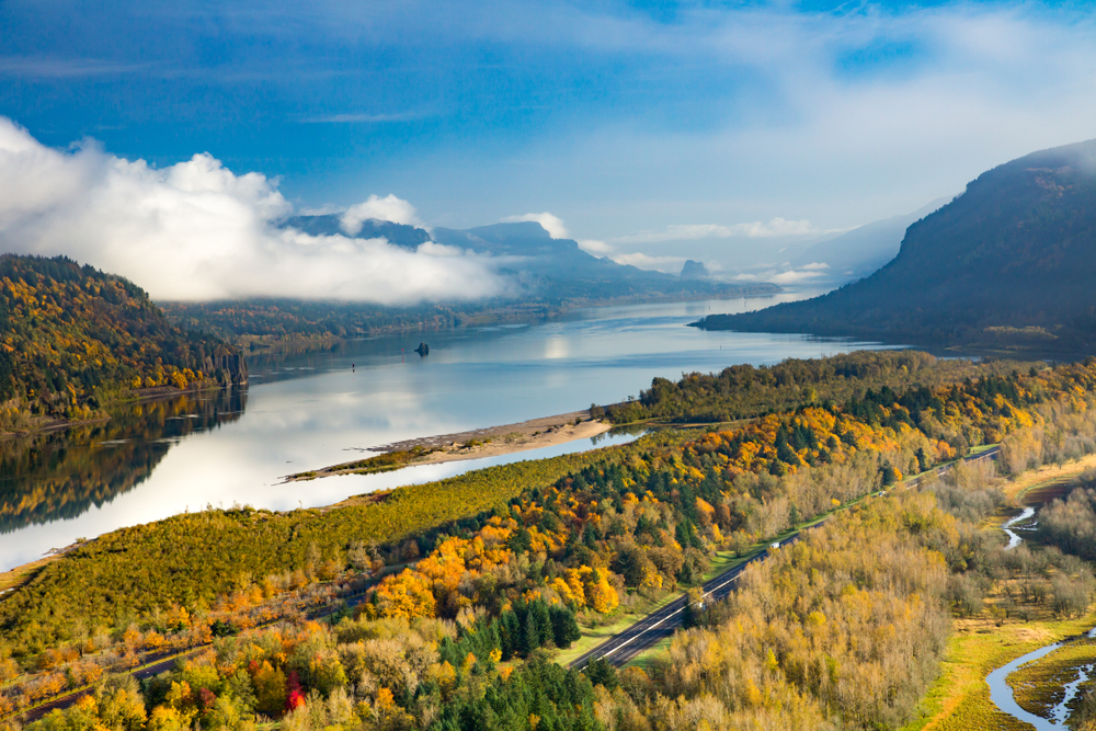 On a road in Oregon, a road curves through yellow trees toward mountains with a lake beyond.