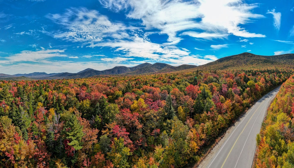 An overhead photo in New Hampshire shows a road stretching on the cusp of trees changing colors, going to mountains.