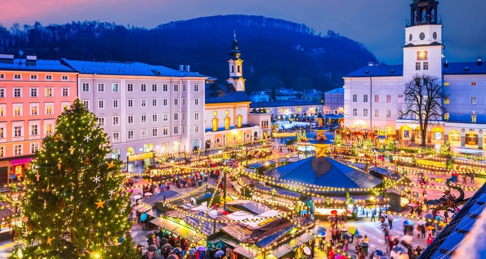 christmas market with bustling people and lights surrounded by buildings. there is a christmas tree off to the left and a hill with trees on it behind the buildings