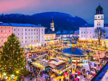 christmas market with bustling people and lights surrounded by buildings. there is a christmas tree off to the left and a hill with trees on it behind the buildings