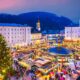 christmas market with bustling people and lights surrounded by buildings. there is a christmas tree off to the left and a hill with trees on it behind the buildings