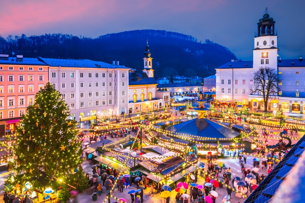 christmas market with bustling people and lights surrounded by buildings. there is a christmas tree off to the left and a hill with trees on it behind the buildings