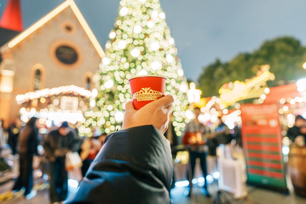 a woman in black jacket is holding a cup of mulled wine in front of a christmas tree in Germany market