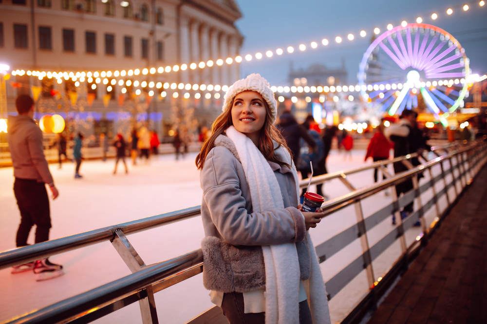 a woman dressed warm standing in front of a carousel and ice skating rink at dusk