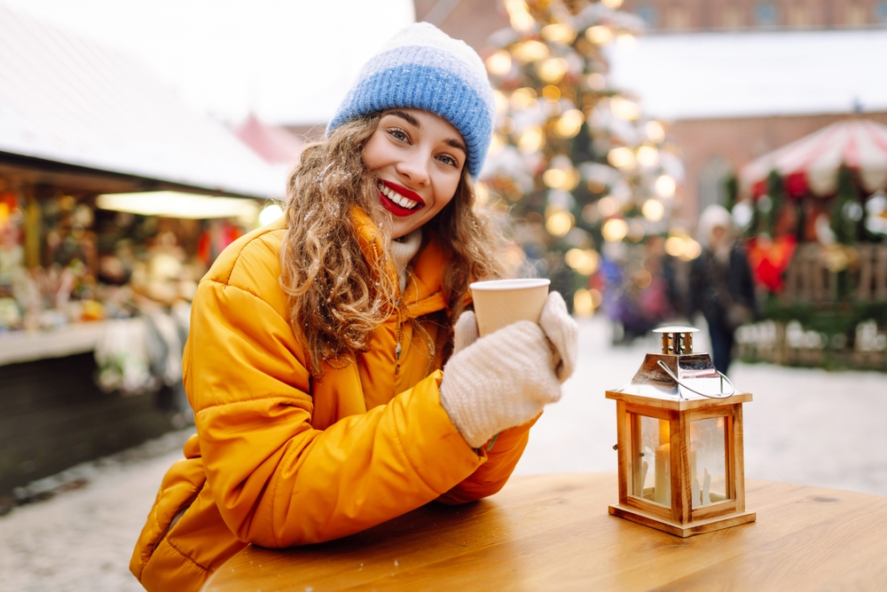 A young girl in red lipstick knows what to wear to Christmas markets in Europe as she stands a table and sips a hot drink while wearing a parka, gloves and hat.