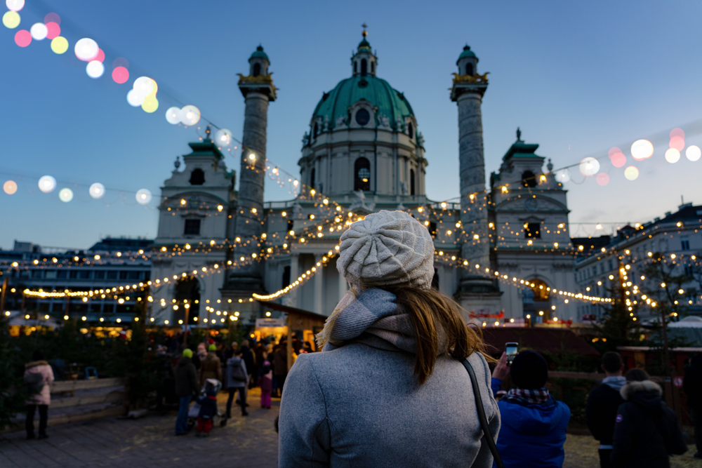 A woman looks up at a stunning building and the lights that highlight the crowd while wearing a scarf, hat and more.