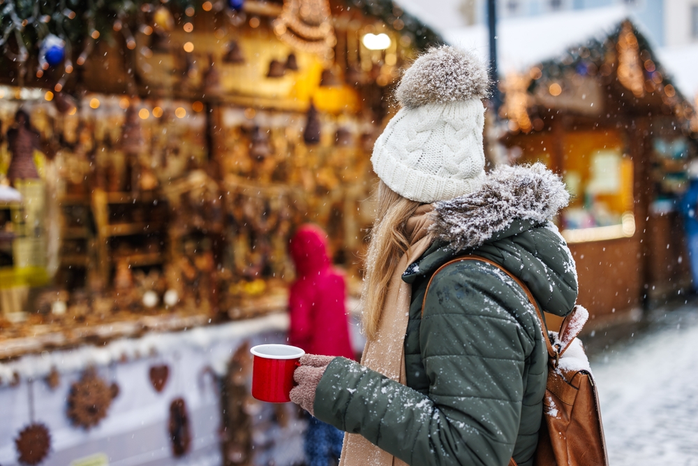 A woman turns to face a market, and she knows what to wear to Christmas markets in Europe in her parka, gloves and hat, as she holds a cup of hot coco.