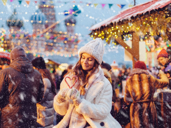 A girl knows what to wear to Christmas markets in Europe as she creates a heart out of candy canes while wearing a knit hat, a chunky coat and more in front of stalls.