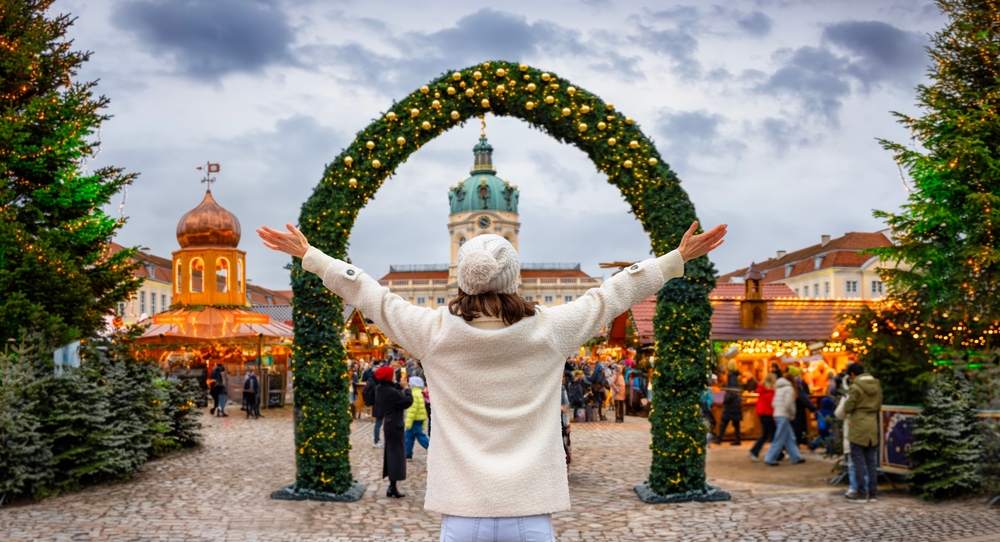 A young girl stands on the cusp of a Christmas market entrance, a green arch decorated beckoning her as she spreads her arms wide in her wool sweater and hat.