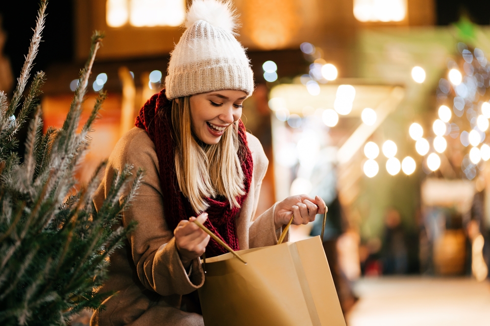 A girl looks into a paper bag excitedly while wearing a scarf, hat and jacket.