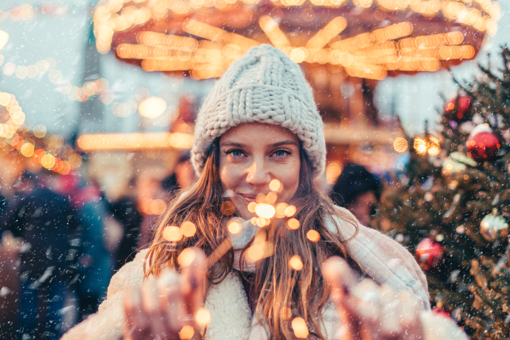 A girl in a knot hat and sweater knows what to wear to Christmas markets in Europe as she stands beyond lights, holding a sparkler at the camera.