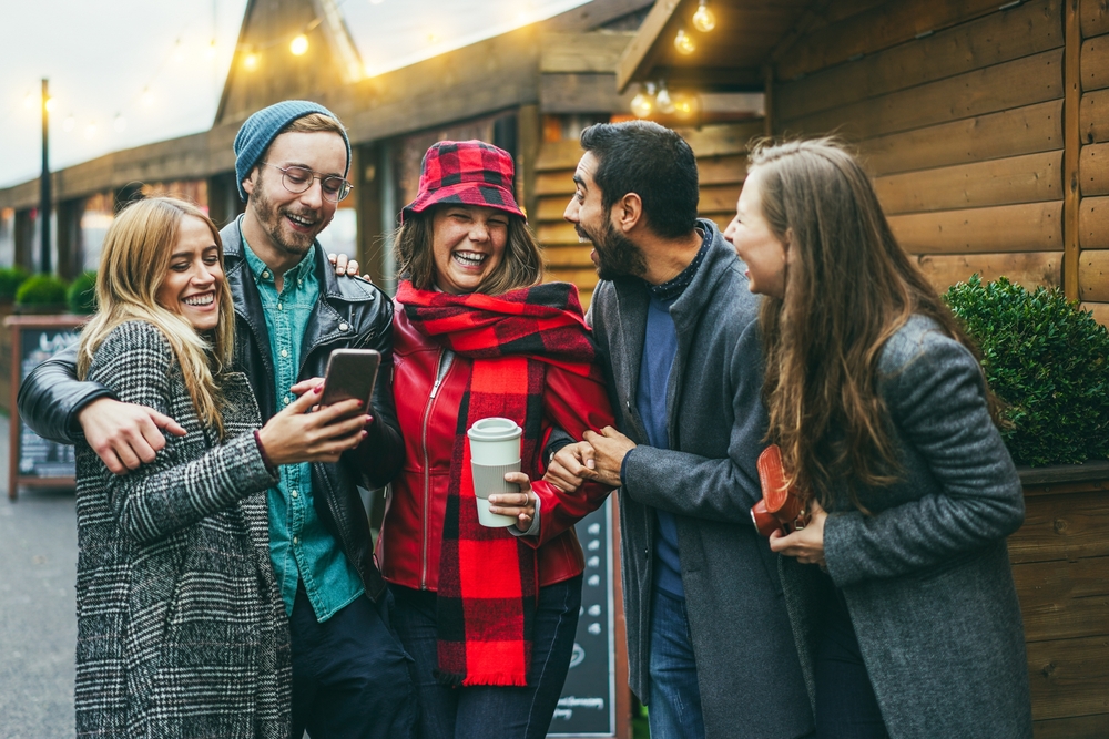 A group of friends gather in front of Christmas stalls, all in long wool cats and hats as they laugh, knowing what to wear to Christmas markets in Europe.