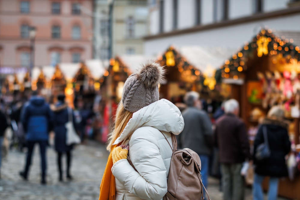A young girl with a backpack turns and looks over her shoulder to see what Christmas market stalls are beyond her, as she knows what to wear to Christmas markets in Europe, with her puffy parka, orange gloves , scarf and knit hat.