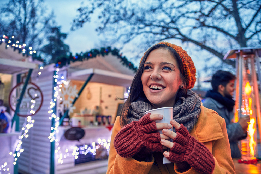 This girl knows what to wear to Christmas markets in Europe as she huddles close to a fire lamp and enjoys a hot drink while wearing a coat, hat and scarf.
