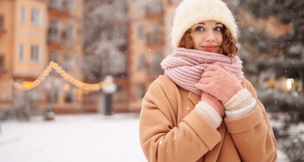As it snows, a woman clutches her knit scarf to her chest, her wool coat, jacket, and gloves keeping her warm.